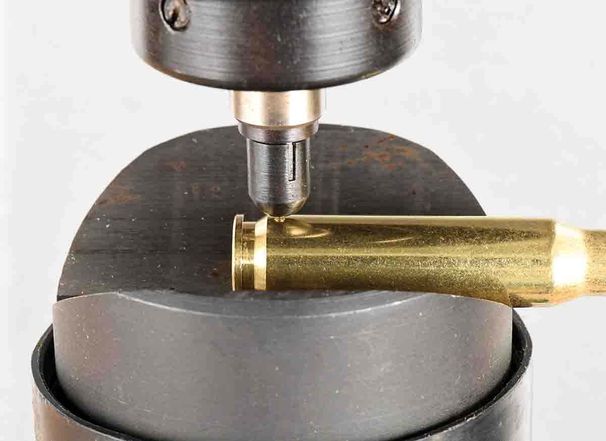 A cartridge case is positioned in the V-block anvil for centering and holding the case while the steel penetrator is pressed into the brass under a specific force. Measurements are taken over the web section just forward of the extractor groove.